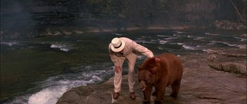 Movie still from “The Jungle Book” (1994), directed by Stephen Sommers – A man standing next to a brown bear near a body of water; Wide shot, High angle