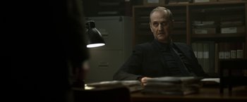 Movie still from “Department Q: The Keeper of Lost Causes” (2013), directed by Mikkel Nørgaard – A man sitting at a table in front of a lamp; Medium shot, Over the shoulder angle