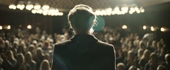 Movie still from “Department Q: The Keeper of Lost Causes” (2013), directed by Mikkel Nørgaard – A person is standing in front of an audience; Medium shot, Over the shoulder angle