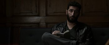 Movie still from “Department Q: The Keeper of Lost Causes” (2013), directed by Mikkel Nørgaard – A man sitting on top of a couch holding an open book; Close Up shot, Over the shoulder angle