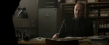 Movie still from “Department Q: The Keeper of Lost Causes” (2013), directed by Mikkel Nørgaard – A man sitting at a desk in a room; Medium shot, Low angle