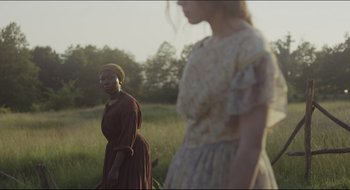 Movie still from “The Keeping Room” (2014), directed by Daniel Barber – Two women standing in a grassy field with trees in the background; Medium shot, Over the shoulder angle