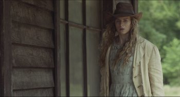 Movie still from “The Keeping Room” (2014), directed by Daniel Barber – A woman in a hat and dress standing in front of a window; Medium shot, Over the shoulder angle