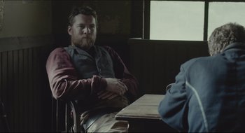 Movie still from “The Keeping Room” (2014), directed by Daniel Barber – A man sitting at a table with his arms crossed; Medium shot, Over the shoulder angle