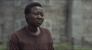 Movie still from “The Keeping Room” (2014), directed by Daniel Barber – A woman in a brown shirt is standing in front of a brick building; Close Up shot, Over the shoulder angle