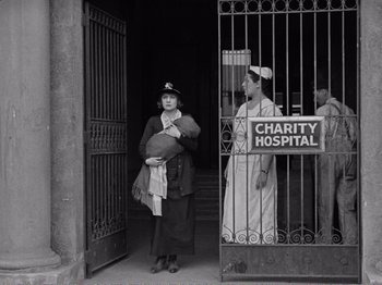 Movie still from “The Kid” (1921), directed by Charles Chaplin – A woman holding a baby standing in front of a hospital; Wide shot, Low angle