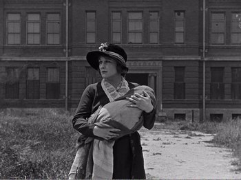 Movie still from “The Kid” (1921), directed by Charles Chaplin – A woman holding a baby in her arms in front of an abandoned building; Wide shot, Low angle