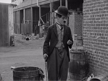 Movie still from “The Kid” (1921), directed by Charles Chaplin – An old photo of a man in a suit and a hat standing in front of a brick building; Wide shot, Over the shoulder angle