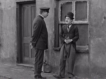 Movie still from “The Kid” (1921), directed by Charles Chaplin – A man in a suit talking to another man in a hat; Medium shot, Over the shoulder angle