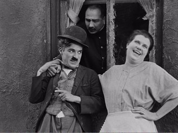 Movie still from “The Kid” (1921), directed by Charles Chaplin – An old black and white photo of a man and two women; Medium shot, Low angle