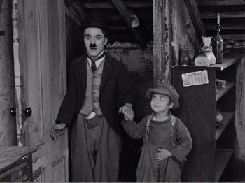 Movie still from “The Kid” (1921), directed by Charles Chaplin – A man and a boy holding hands in front of an open door; Medium shot, High angle