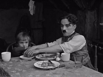 Movie still from “The Kid” (1921), directed by Charles Chaplin – A man sitting at a dinner table with a child; Medium shot, High angle