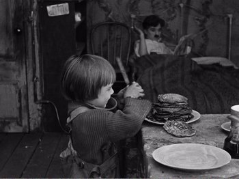 Movie still from “The Kid” (1921), directed by Charles Chaplin – A young boy sitting at a table with a plate of pancakes on it; Medium shot, High angle