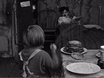 Movie still from “The Kid” (1921), directed by Charles Chaplin – A woman sitting at a table with a plate of pancakes in front of a man; Medium shot, Overhead angle