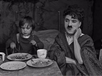 Movie still from “The Kid” (1921), directed by Charles Chaplin – A man and a boy sitting at a table with plates of food on it; Medium shot, High angle