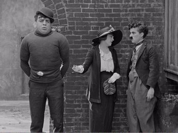 Movie still from “The Kid” (1921), directed by Charles Chaplin – An old photo of a man , woman , and a man standing next to a brick wall; Medium shot, Over the shoulder angle