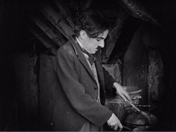 Movie still from “The Kid” (1921), directed by Charles Chaplin – A man in a jacket and tie cooking food in an oven in an attic; Medium shot, Low angle