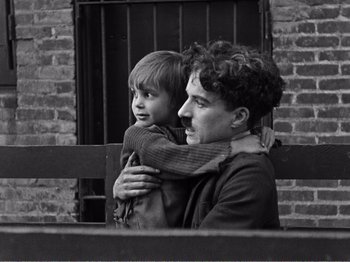 Movie still from “The Kid” (1921), directed by Charles Chaplin – A man and a boy are standing next to a brick wall; Medium shot, High angle