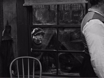 Movie still from “The Kid” (1921), directed by Charles Chaplin – A young boy wearing a hat looking out of a window; Wide shot, High angle