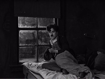 Movie still from “The Kid” (1921), directed by Charles Chaplin – A man sitting on top of a bed next to a window; Medium shot, High angle