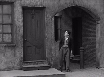 Movie still from “The Kid” (1921), directed by Charles Chaplin – An old photo of a man standing in front of a door; Wide shot, Low angle
