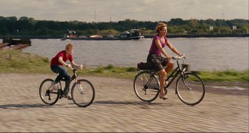 Movie still from “The Kid with a Bike” (2011), directed by Jean-Pierre Dardenne – Two people riding bikes on a path near a body of water; Wide shot, High angle