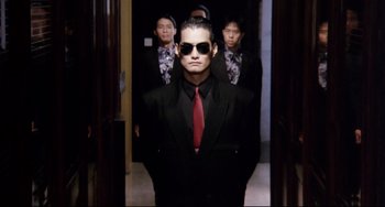 Movie still from “The Killer” (1989), directed by John Woo – A man in a suit and tie standing in front of a group of people; Medium shot, Low angle