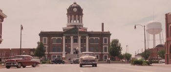Movie still from “The Killer Inside Me” (2010), directed by Michael Winterbottom – An old car is driving past a building with a clock tower; Extreme Wide shot, High angle
