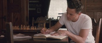 Movie still from “The Killer Inside Me” (2010), directed by Michael Winterbottom – A man writing in a notebook on a wooden table; Medium shot, High angle
