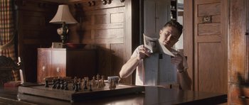 Movie still from “The Killer Inside Me” (2010), directed by Michael Winterbottom – A man reading a newspaper while sitting in front of a chessboard; Medium shot, Low angle