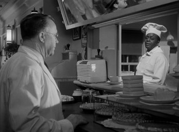 Movie still from “The Killers” (1946), directed by Robert Siodmak – A black and white photo of a man and a woman in a kitchen; Medium shot, Over the shoulder angle