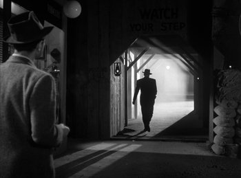 Movie still from “The Killers” (1946), directed by Robert Siodmak – A man in a hat and a suit walking through a tunnel; Wide shot, Over the shoulder angle