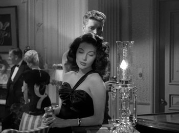 Movie still from “The Killers” (1946), directed by Robert Siodmak – A woman in a black dress holding a wine glass in front of a man; Medium shot, Over the shoulder angle