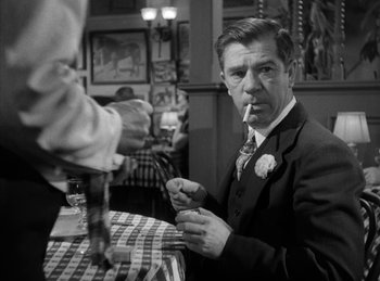 Movie still from “The Killers” (1946), directed by Robert Siodmak – A black and white photo of a man smoking a cigarette at a table; Medium shot, Over the shoulder angle