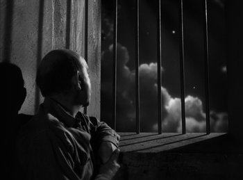 Movie still from “The Killers” (1946), directed by Robert Siodmak – A man standing in front of a window with a cloudy sky behind him; Wide shot, Low angle