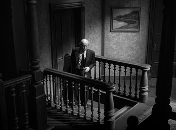 Movie still from “The Killers” (1946), directed by Robert Siodmak – A man in a suit sitting on top of a wooden stair railing; Wide shot, High angle