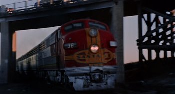 Movie still from “The Killers” (1964), directed by Don Siegel – A red and yellow train traveling down train tracks under a bridge; Extreme Wide shot, Low angle