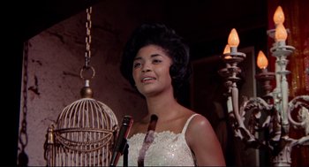 Movie still from “The Killers” (1964), directed by Don Siegel – A woman in a white dress standing next to a bird cage; Close Up shot, Low angle