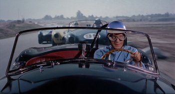 Movie still from “The Killers” (1964), directed by Don Siegel – A man driving a vintage race car in a field; Medium shot, Over the shoulder angle