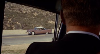 Movie still from “The Killers” (1964), directed by Don Siegel – A man in a suit driving a car down a street; Wide shot, Over the shoulder angle