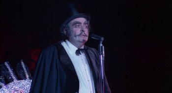 Movie still from “The Killing of a Chinese Bookie” (1976), directed by John Cassavetes – A man with a mustache wearing a top hat and holding a microphone in front of him; Close Up shot, Low angle