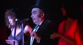 Movie still from “The Killing of a Chinese Bookie” (1976), directed by John Cassavetes – A man in a black suit and a black hat singing; Close Up shot, Low angle