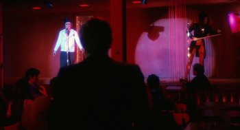 Movie still from “The Killing of a Chinese Bookie” (1976), directed by John Cassavetes – A man standing in front of a screen with a microphone; Wide shot, Over the shoulder angle