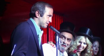 Movie still from “The Killing of a Chinese Bookie” (1976), directed by John Cassavetes – A man standing next to another man wearing a top hat and clown makeup; Medium shot, Over the shoulder angle