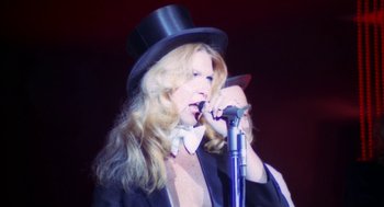 Movie still from “The Killing of a Chinese Bookie” (1976), directed by John Cassavetes – A woman wearing a top hat while holding a microphone; Medium shot, Low angle