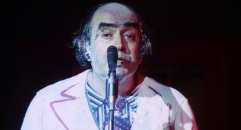 Movie still from “The Killing of a Chinese Bookie” (1976), directed by John Cassavetes – A man in a white jacket is speaking into a microphone; Close Up shot, Low angle