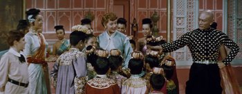 Movie still from “The King and I” (1956), directed by Walter Lang – A group of people standing around each other in front of a building; Medium shot, Over the shoulder angle
