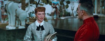 Movie still from “The King and I” (1956), directed by Walter Lang – A woman holding a book while standing in front of a painting; Medium shot, Low angle