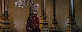 Movie still from “The King and I” (1956), directed by Walter Lang – A bald man in a red and gold outfit stands in front of a wall; Medium shot, Low angle