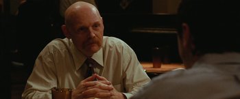 Movie still from “The King of Staten Island” (2020), directed by Judd Apatow – A bald man with a mustache sitting at a table with his hands crossed; Close Up shot, Over the shoulder angle